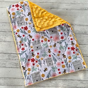 May include: A soft baby blanket featuring a white background with a repeating pattern of gray beehives, yellow honeycombs, small bees, and colorful red, pink, and yellow flowers. The reverse side is a solid, textured yellow minky fabric.
