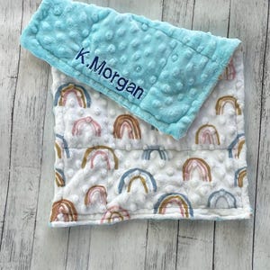 May include: A baby blanket with a light blue, textured top and a white base. The base features a rainbow pattern in pastel colors. The name "K.Morgan" is embroidered in blue on the top section. The blanket is designed for comfort.