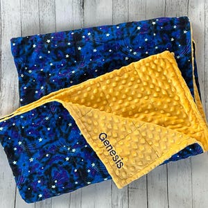 May include: A folded blanket featuring a dark blue outer side with a white star and galaxy pattern. The inner side is a solid mustard yellow fabric with a raised dimpled texture. The name Genesis is embroidered in black on the yellow side.
