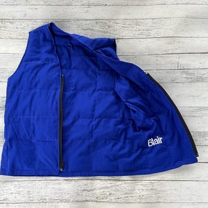 May include: A vibrant blue vest with a quilted pattern and a black zipper closure. The vest is open, revealing the interior lining and the embroidered name "Blair" in white. The vest is made of a soft, possibly padded material.