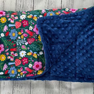 May include: A rectangular blanket featuring a dark green background with a colorful pattern of pink flowers, red mushrooms, yellow butterflies, and green leaves. The reverse side is a solid navy blue fabric with a raised, textured dimpled surface.
