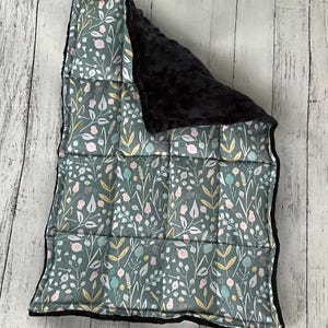 May include: A rectangular quilted blanket featuring a sage green background with a repeating pattern of white, pink, and yellow botanical illustrations including leaves and flowers. The reverse side is a solid black textured fabric with a dimpled surface.