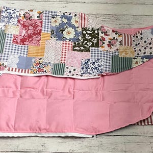 May include: A quilted fabric item featuring a patchwork top layer with various floral, gingham, and striped patterns in shades of red, blue, green, and yellow. The underside is a solid light pink fabric, and a white zipper runs along the edge.