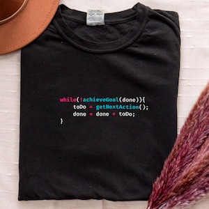May include: Black t-shirt with a programming code snippet in white, pink, and teal. The code reads "while(!achieveGoal(done)) { toDo = getNextAction(); done = done + toDo; }." A brown hat and decorative dried flowers are in the background.