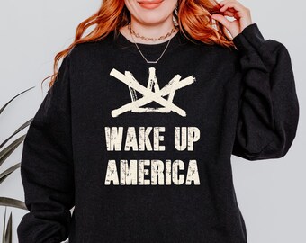 May include: Dark gray sweatshirt with the text "WAKE UP AMERICA" in a distressed, off-white font. Above the text is a white, abstract design. The sweatshirt has long sleeves and a crew neck.