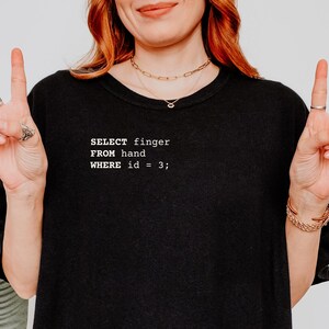 May include: Black t-shirt with white text that reads "SELECT finger FROM hand WHERE id = 3;". The person is wearing gold jewelry and making a peace sign with their fingers.