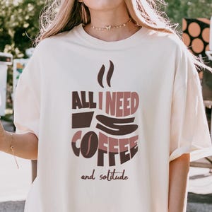 May include: A white t-shirt with a brown and pink graphic of a coffee cup and the text "All I need is coffee and solitude".