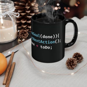 May include: Black ceramic mug with the text "WeGoal(done){etNextAction(); one + toDo;" in a computer code style. The mug is filled with a dark liquid, possibly coffee or tea, and is surrounded by decorative items.