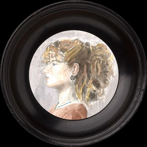 May include: A watercolour portrait of a woman in profile, framed in a round, black wooden frame. The woman has light skin, brown hair, and wears a brown top and necklace. The background is a soft grey. The frame is approximately 15 cm in diameter.