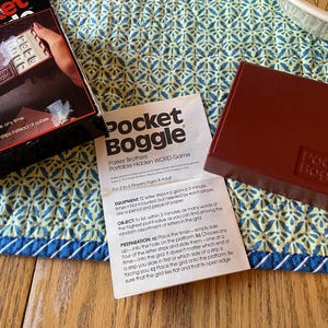 May include: A vintage Pocket Boggle game by Parker Brothers. The image shows the game box, instructions, and a maroon game case. The box features the game name and a picture of the game in use. The instructions are open, and the game case is closed.