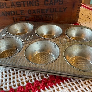 May include: A vintage, well-worn, six-cup muffin tin made of silver-colored metal. The tin has a rectangular shape with rounded cups and a diamond pattern at the bottom of each cup. The text "BLASTING CAPS HANDLE CAREFULLY" is visible in the background.