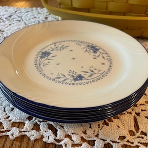 May include: A stack of round white ceramic plates featuring a blue floral wreath pattern in the center and a dark blue rim. The plates have a subtle embossed swirl design on the outer edge, creating a textured appearance on the surface.