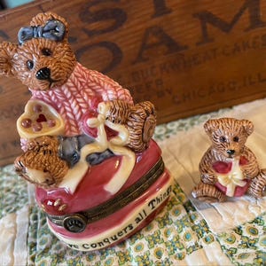 May include: Two ceramic teddy bear figurines. One is a jewelry box with a pink sweater, blue bow, and a heart-shaped lid. The other is a smaller bear holding a gift. The box reads "Love Conquers all Things."