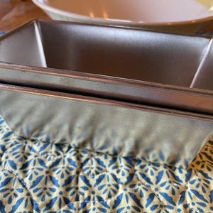 May include: Two rectangular, silver-colored loaf pans stacked on top of each other. The pans have a slightly trapezoidal shape with a copper-colored rim. They sit on a blue and yellow floral patterned quilted surface. A white and yellow bowl is in the background.