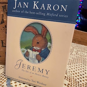 May include: A hardcover book titled "Jeremy: The Tale of an Honest Bunny" by Jan Karon. The cover features an illustration of a bunny in a blue jacket reading a paper. The book has a blue ribbon bookmark.