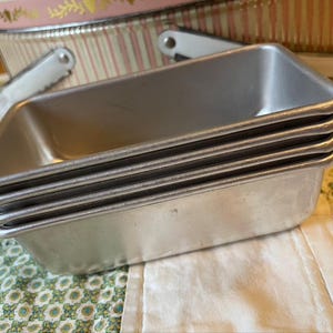 May include: A stack of four rectangular, silver-colored loaf pans. The pans are made of metal and have a slightly rounded edge. In the background, a pink and white striped tin with a handle is visible, suggesting a vintage or retro theme.