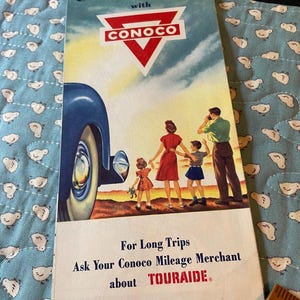 May include: A vintage Conoco travel brochure featuring a blue car and a family of four standing in a landscape. The text reads: Travel United States with CONOCO, For Long Trips Ask Your Conoco Mileage Merchant about TOURAIDE. A wooden ruler is visible.