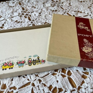 May include: A vintage stationery set featuring a box with a cream-colored exterior and a red stripe with floral illustrations. The open box reveals stationery with a "Happy Birthday" greeting and a train illustration with floral accents.