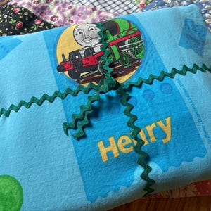 May include: A light blue pillowcase featuring a cartoon train character with the name "Henry" in yellow. The pillowcase is tied with a green zigzag ribbon. The pillowcase is on a patchwork quilt.