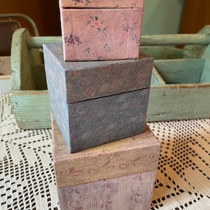 May include: A stack of three square boxes. The top box is pink with a small floral pattern. The middle box is blue-gray with a geometric diamond and floral print. The bottom box is light purple and beige with a vertical striped and scroll pattern.