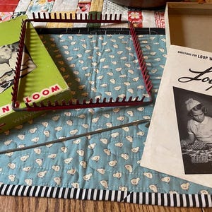 May include: A vintage Loop Craft weaving loom set including a green cardboard box with black and white illustrations, a red metal square loom frame with pegs, a long metal weaving needle, and an instructional booklet titled Loop Craft.