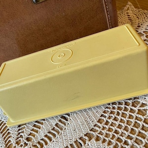 May include: A vintage, rectangular, yellow Tupperware container. The lid has the Tupperware logo and text. The container is on a lace doily, with a brown leather case in the background.