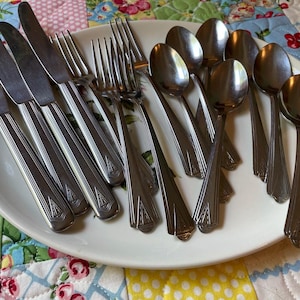 May include: A collection of stainless steel flatware including four dinner knives, three forks, and five spoons arranged on a white oval platter. Each piece features a polished finish and a geometric Art Deco style embossed pattern on the handle.