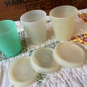 May include: Three translucent plastic cups in mint green, off-white, and pale yellow, accompanied by three matching circular off-white lids. Each cup features horizontal ridges along the exterior surface, and the lids are embossed with the Tupperware brand name.