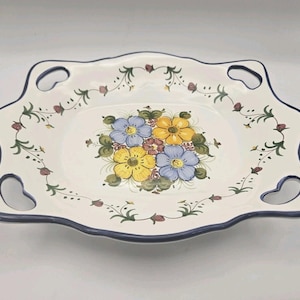 Puede incluir: Plato de cerámica blanco con borde festoneado y cuatro asas en forma de corazón. El plato presenta un diseño floral en el centro con flores amarillas, azules y moradas. Un borde azul y detalles de vid floral completan el diseño.