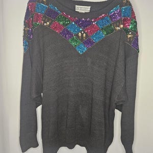 Pode incluir: Um suéter cinza com um design de lantejoulas na gola. As lantejoulas são de várias cores, incluindo verde, azul, rosa e roxo. O suéter tem uma gola redonda e mangas compridas. O rótulo no suéter diz "PK Elliot".