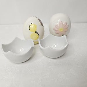 May include: A set of four ceramic Easter-themed items. Two egg-shaped ornaments are decorated with a yellow butterfly and a pink flower. Two white, eggshell-shaped bowls are also included.