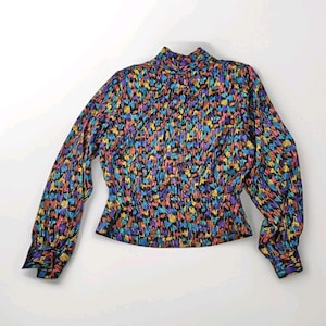Op de afbeelding: Een vintage blouse met een hoge halslijn en lange mouwen. De blouse heeft een kleurrijk, abstract patroon in tinten blauw, paars, oranje en geel op een zwarte achtergrond. De mouwen hebben manchetten met knopen en de taille is getailleerd.
