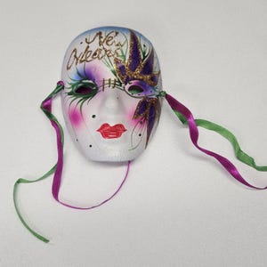Peut inclure: Un masque décoratif avec une base blanche, représentant un visage peint avec des lèvres rouges, des joues roses et des accents d'yeux verts. Le masque est orné d'ornements violets et dorés et du texte "New Orleans". Des rubans violets et verts sont attachés.