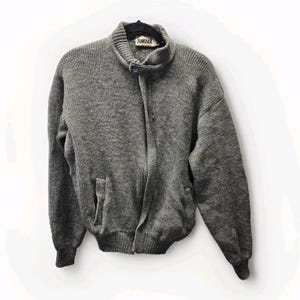 May include: A gray, knitted zip-up jacket with a high collar and a small pocket on the left side. The jacket has a ribbed hem and cuffs. The brand name "TUNDRA" is visible on a tag near the collar. The jacket is made of a thick, warm material.