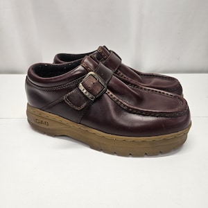 Può includere: Paio di scarpe in pelle bordeaux con chiusura a fibbia e suola color cuoio. Le scarpe hanno la punta arrotondata e un tacco basso. La fibbia riporta le lettere "SAO".