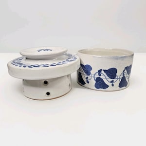 May include: A two-piece ceramic butter dish, featuring a white base and lid decorated with blue leaf patterns. The lid has a small handle and a matching leaf design. The base has several small holes. A matching ceramic container with blue leaf patterns is also present.