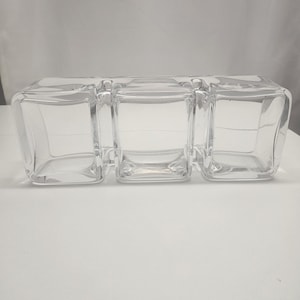 Può includere: Un piatto da portata in vetro trasparente con tre scomparti quadrati. Il piatto è realizzato in vetro spesso e ha un design semplice e moderno. I scomparti sono collegati e poggiano su una superficie bianca.