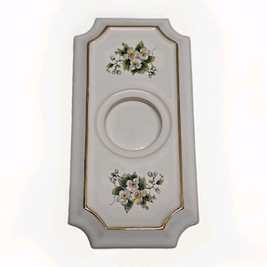 May include: A rectangular white ceramic wall plate featuring a central circular opening. The plate is decorated with two floral clusters of white blossoms and green leaves, one above and one below the center. A thin gold trim outlines the inner panel.