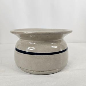 May include: A beige ceramic butter dish with a dark blue horizontal stripe. The dish has a rounded body and a flared rim. The butter dish is a classic design, suitable for kitchen decor.