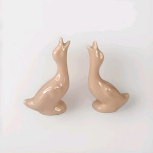 May include: Two ceramic goose ornaments in a soft, muted pink colour. The geese have elongated necks and open beaks, giving them a stylized, elegant appearance. These decorative items are ideal for home decor.