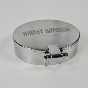May include: A shiny, chrome-colored, round metal container with the words "HARLEY-DAVIDSON" engraved on the lid. The container has a hinged clasp and a polished finish, reflecting the light. The product is likely a small storage box or accessory.