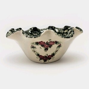 May include: Cream ceramic bowl with a ruffled edge and a heart-shaped floral design. The rim has a black and white speckled pattern. The heart is formed by green leaves and red roses. A decorative piece for the home.