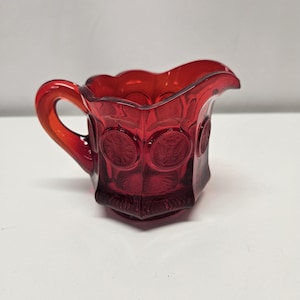 May include: A ruby red glass creamer with a handle and a decorative pattern. The creamer has a scalloped rim and circular embossed designs on the sides. The handle is curved and the glass is transparent.