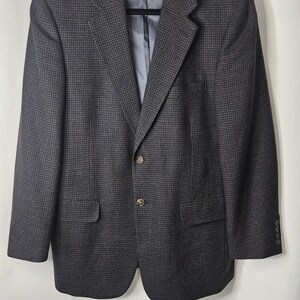 May include: A dark grey houndstooth blazer with a two-button fastening and a notched lapel. The jacket has two front pockets and a chest pocket. The interior lining is a light blue colour. The blazer is hanging on a black hanger.