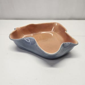 May include: A ceramic serving dish with a wavy rim. The exterior is a muted blue, while the interior has a warm, peach-colored glaze. The dish has a rectangular shape with gently curved sides.