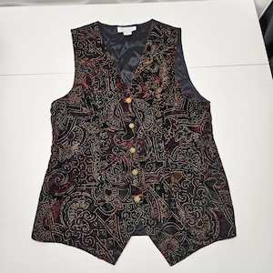 May include: A black velvet vest with an ornate paisley pattern in burgundy, gold, and green. The vest has a V-neck, gold buttons, and a quilted black lining. A vintage-style garment.