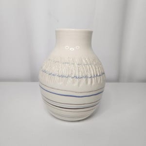 Peut inclure: Un vase en céramique crème avec une section supérieure texturée et des rayures horizontales bleues et marron. Le vase a un corps arrondi et un col étroit, adapté pour exposer des fleurs ou comme pièce décorative.