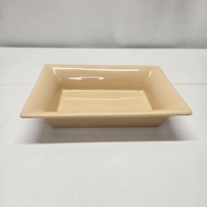 May include: A rectangular, light beige ceramic dish with slightly flared sides. The dish has a smooth, glossy finish and is designed for serving or display. The corners are gently rounded, and the overall shape is modern and minimalist.