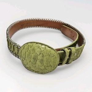 May include: A green belt with a textured, oval buckle. The belt has a snakeskin pattern and a silver-colored trim along the edges. The buckle is centered on the belt, and the leather is a light green color.