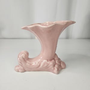 May include: A light pink ceramic vase with a distinctive horn-like form. The vase features a flared top and a curved, ornate base with a textured design. The overall aesthetic is vintage and decorative.